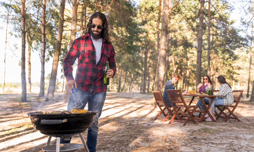 Man grilling food outdoors while friends enjoy a meal in the background